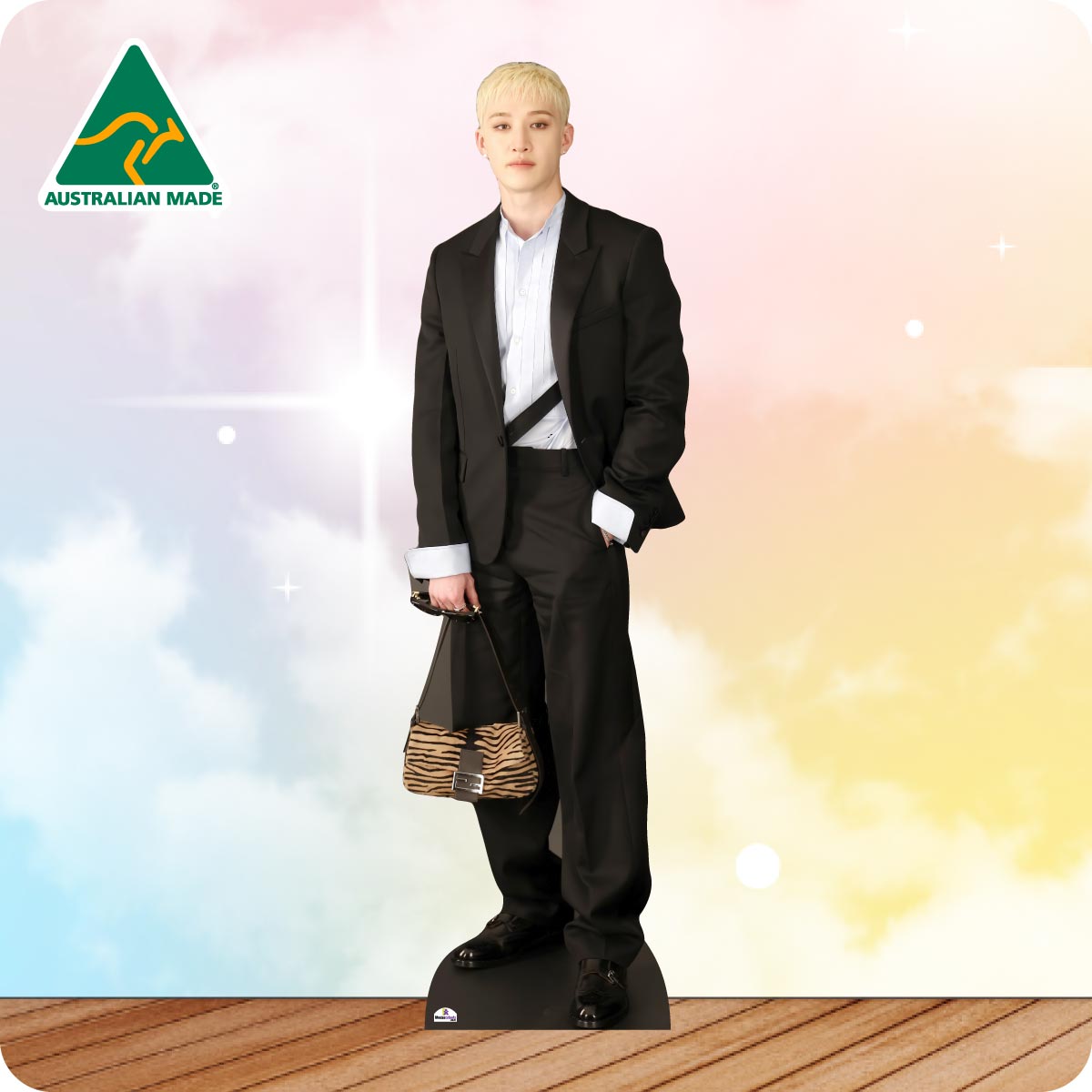A full-body cardboard cutout standee of a young man with short blond hair, wearing a black suit, white shirt, and black belt, holding a zebra-print handbag. The standee is set against a pastel sky background with clouds and a wooden floor, with an 'AUSTRALIAN MADE' logo in the top-left corner.