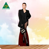 A woman stands in a studio setting wearing a black pinstripe blazer over a matching black pinstripe dress with a high slit, holding a black handbag with a large white monogram. The background is a soft pastel sky gradient with clouds and a wooden floor, plus an 'Australian Made' logo in the top left.