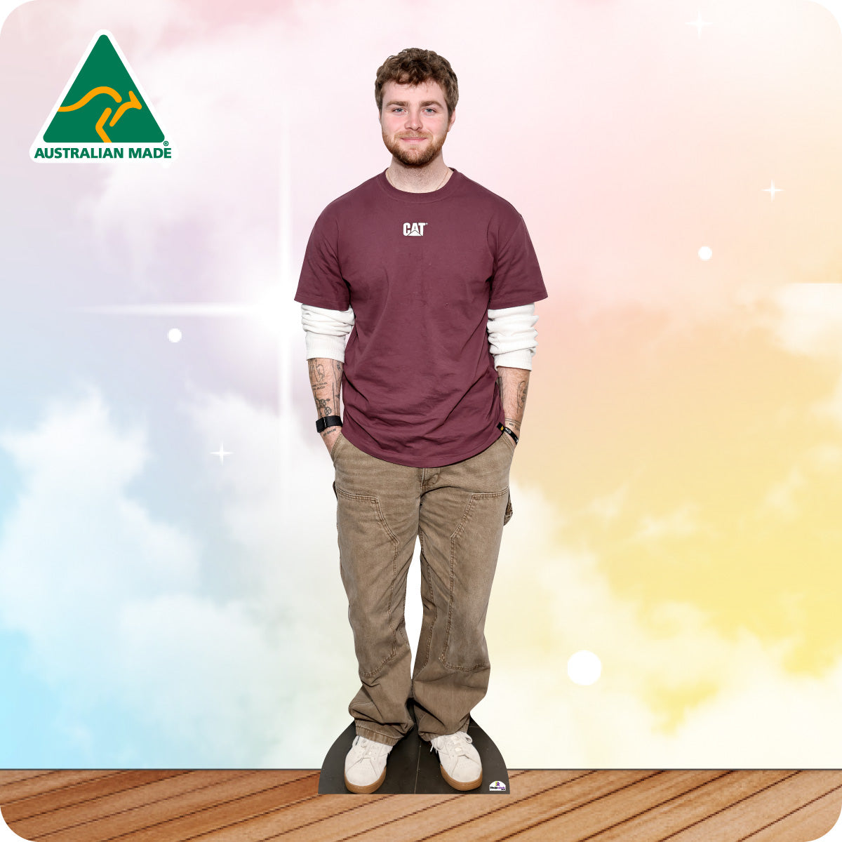 Man wearing a maroon shirt with a logo and brown pants standing on a standee against a colorful gradient background.