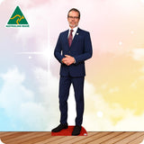 A full-body cardboard cutout of a man in a navy blue suit, white shirt, and burgundy tie, standing on a red base. The background is a pastel sky with clouds in blue, pink, and yellow, with a wooden floor at the bottom.